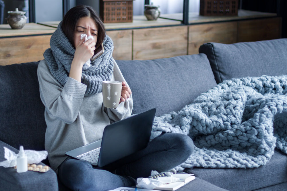 Sick woman blowing nose on couch holding a mug